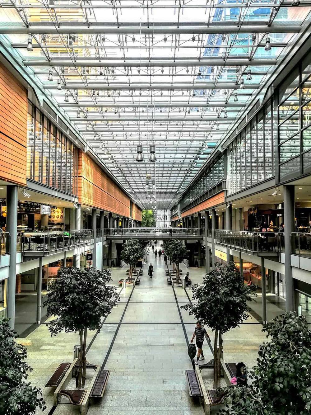 Bright indoor shopping mall with glass ceiling and multiple levels, featuring retail stores and seating areas.