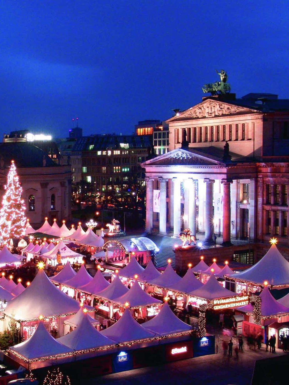 Festive Christmas market in front of historic city hall with illuminated tents and decorated Christmas tree, evening scene in urban holiday setting.