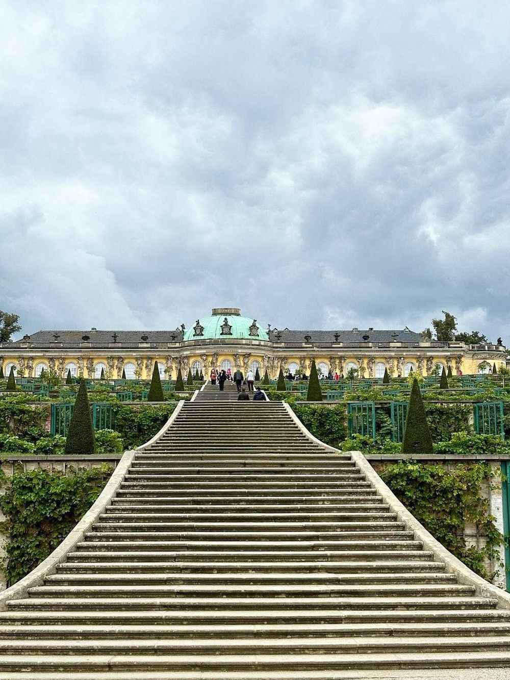 Majestic palace with grand staircase leading to ornate entrance, lush gardens, and cloudy sky in the background.