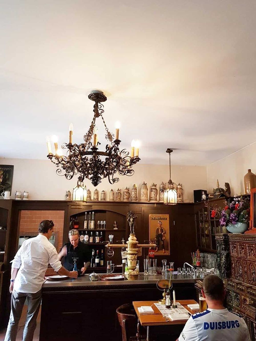 Elegant chandelier hanging above cozy restaurant bar with patrons and staff enjoying dining experience.