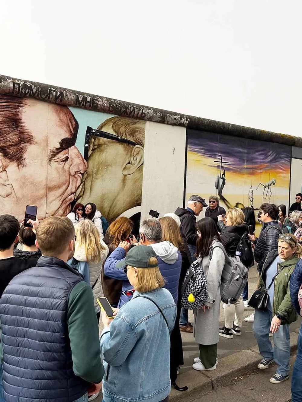Vivid mural art of two men kissing on a large wall, with a crowd gathered for an outdoor event.