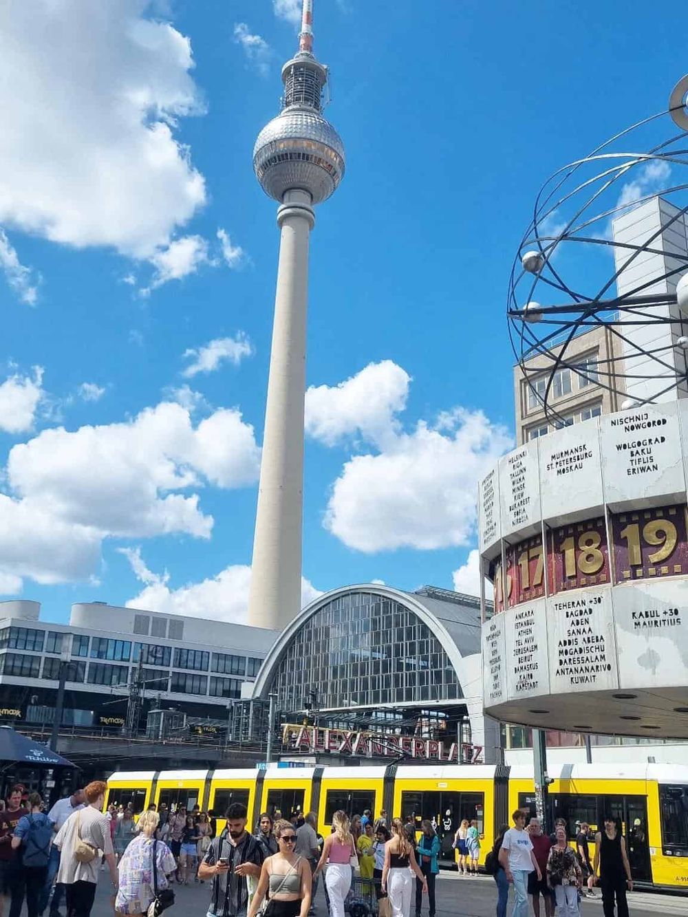 Eiffel Tower with Berlin TV Tower in background, busy city square, crowded with travelers and a yellow tram.