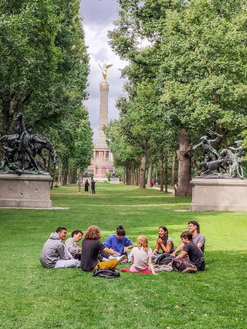 Group of young friends sitting on grass in a park with Berlin Victory Column in background, ideal for exploring Berlin sightseeing.