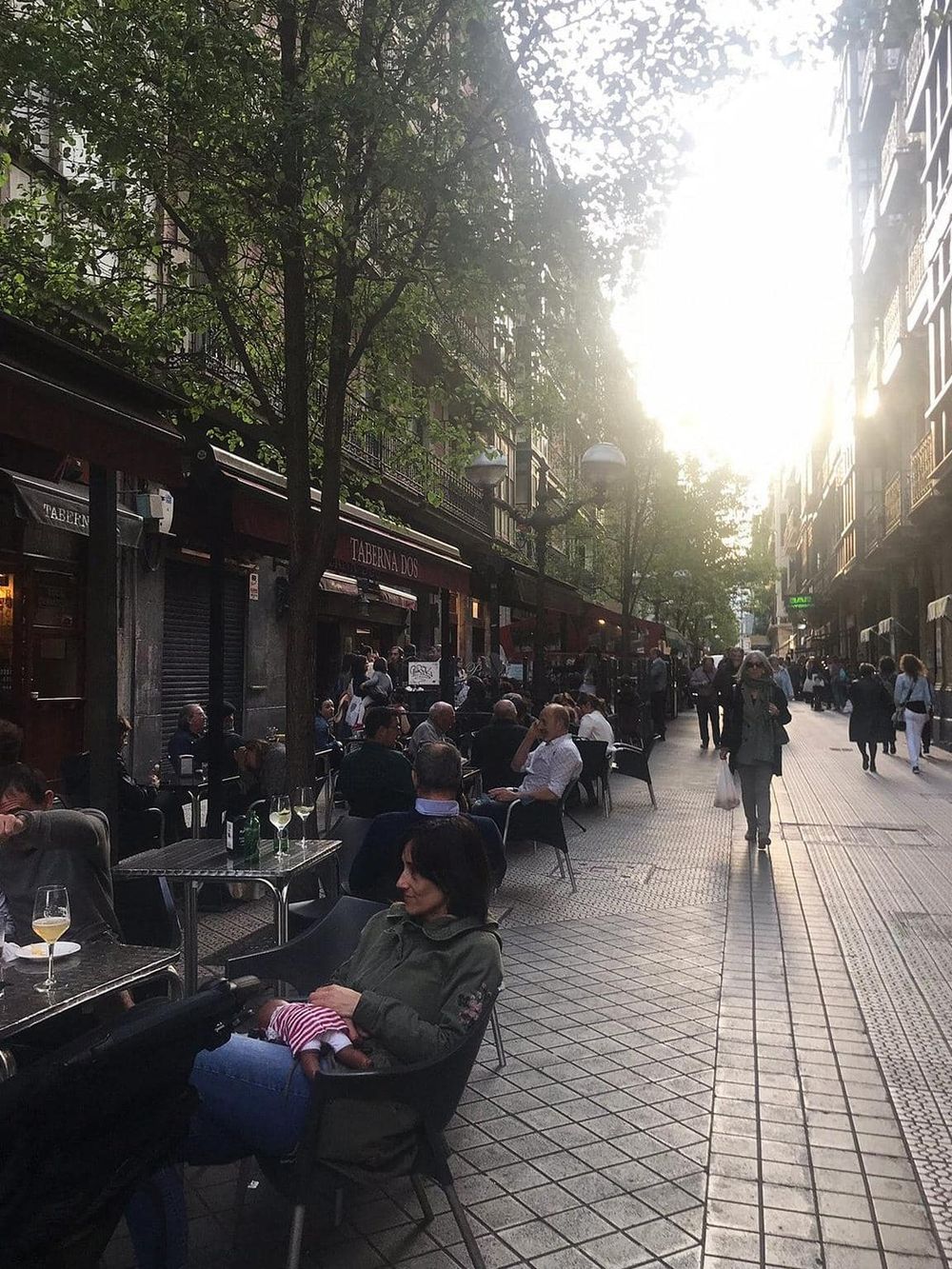Outdoor city street café scene with people dining and walking, sunset lighting, in a bustling urban neighborhood.