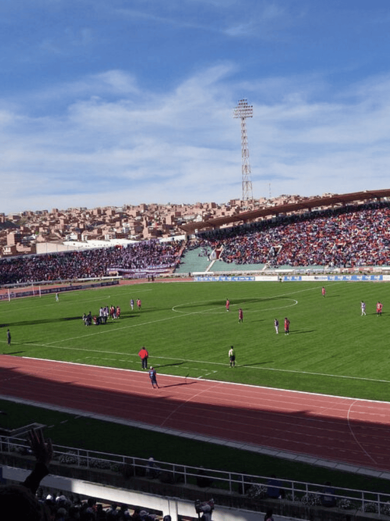 Aerial view of a football stadium with players on the field and packed spectators in the stands.