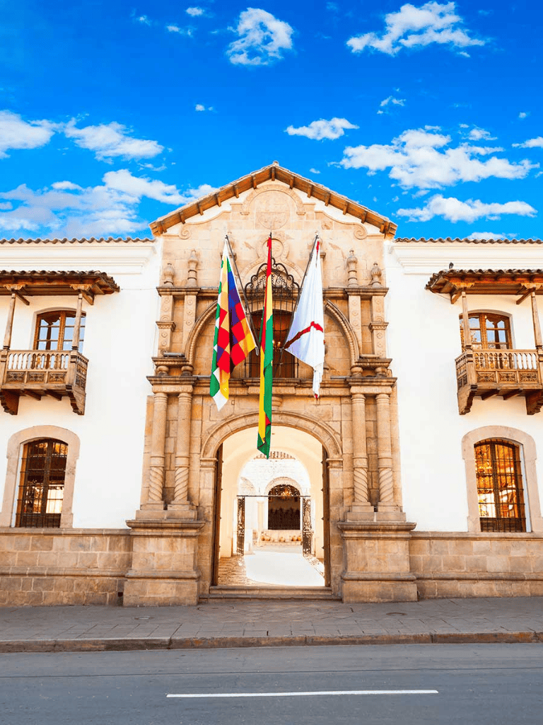 Colorful flags hang outside historic Spanish-style building under bright blue sky.