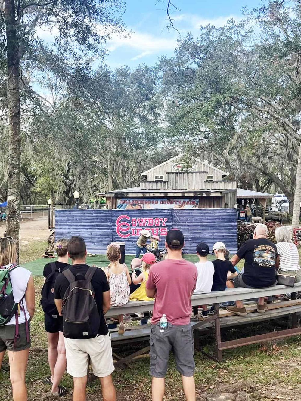 Vibrant outdoor Western-themed cowboy circus performance at county fair with audience and rustic wooden backdrop.