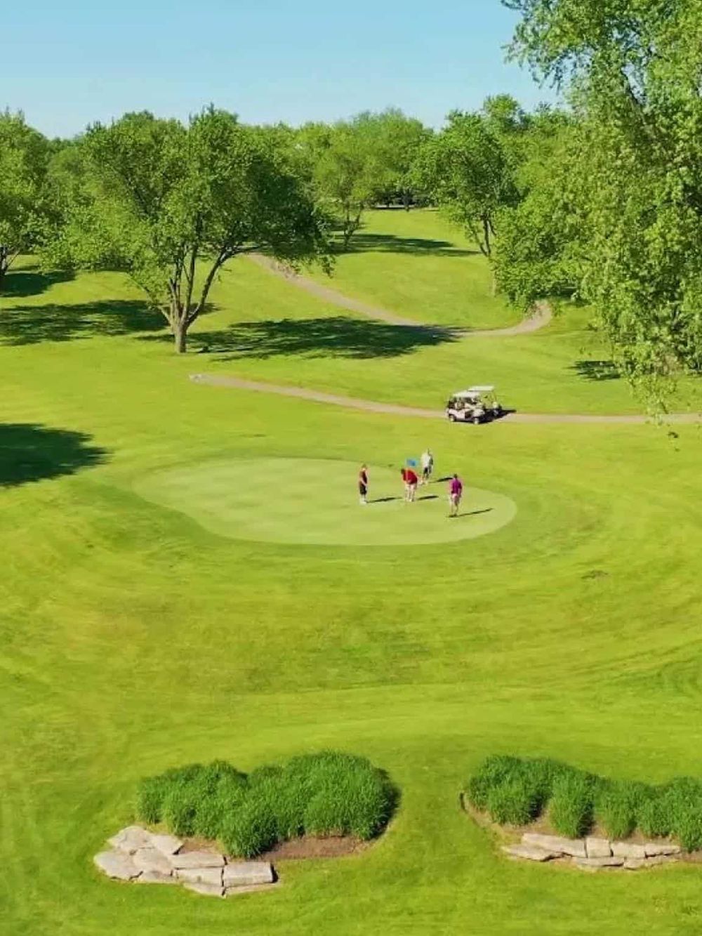 Lush green golf course with players preparing for a game, surrounded by tall trees and scenic landscaping.