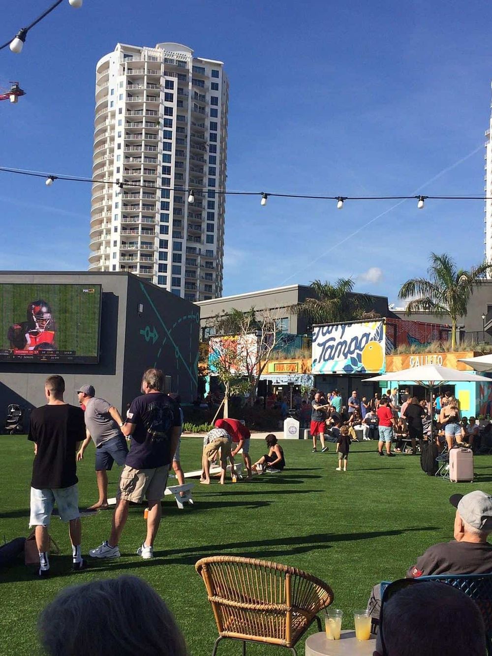 Bright outdoor scene at a local entertainment and dining venue with a large screen, modern buildings, and people enjoying social activities.