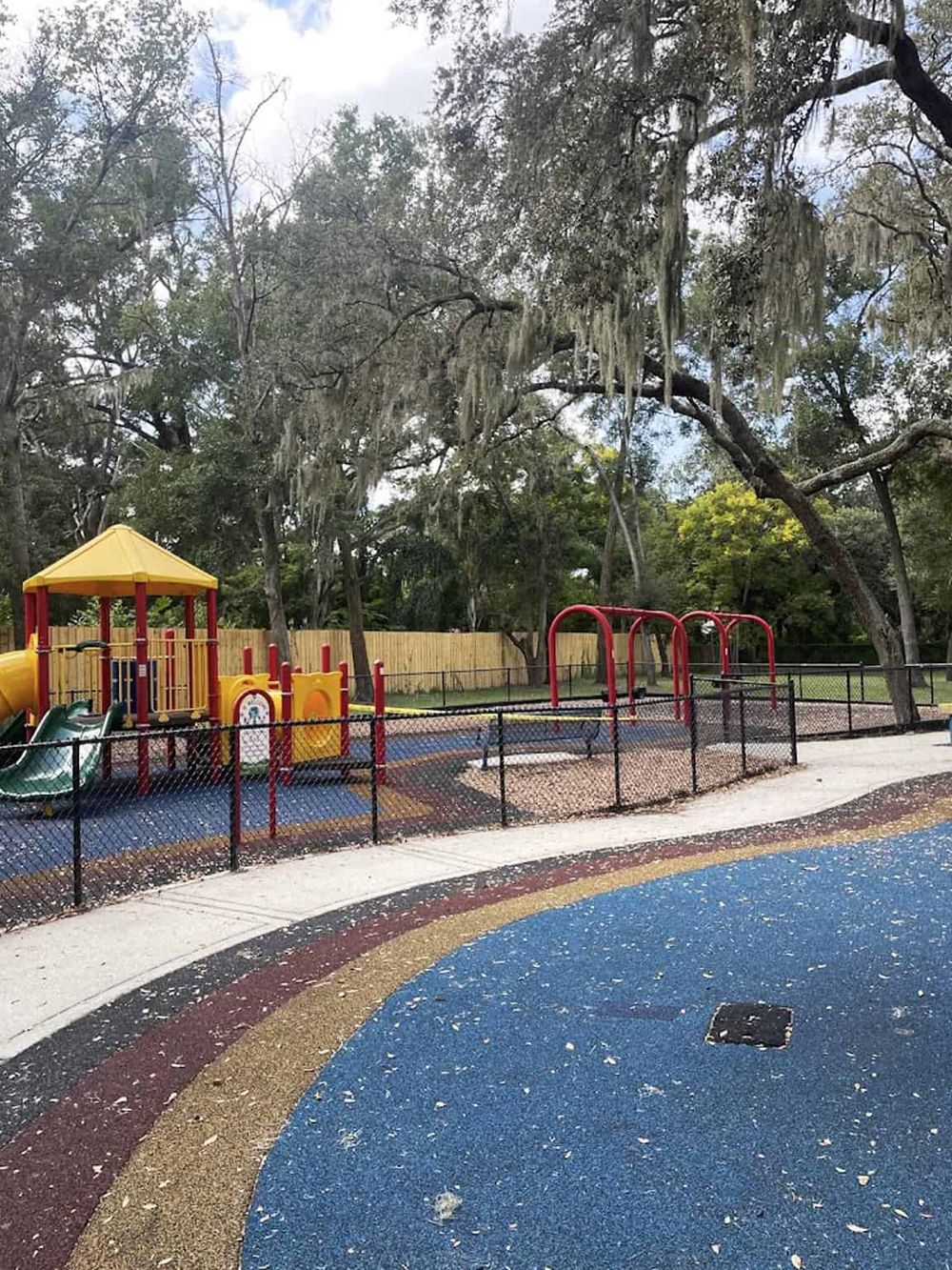 Colorful children's playground with slides and swings in a park setting, surrounded by tall trees and safety fencing.