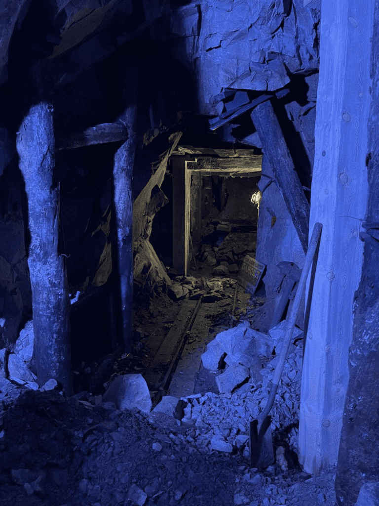 Underground mine tunnel with wooden supports and rocky debris, illuminated in blue light for safety inspections.