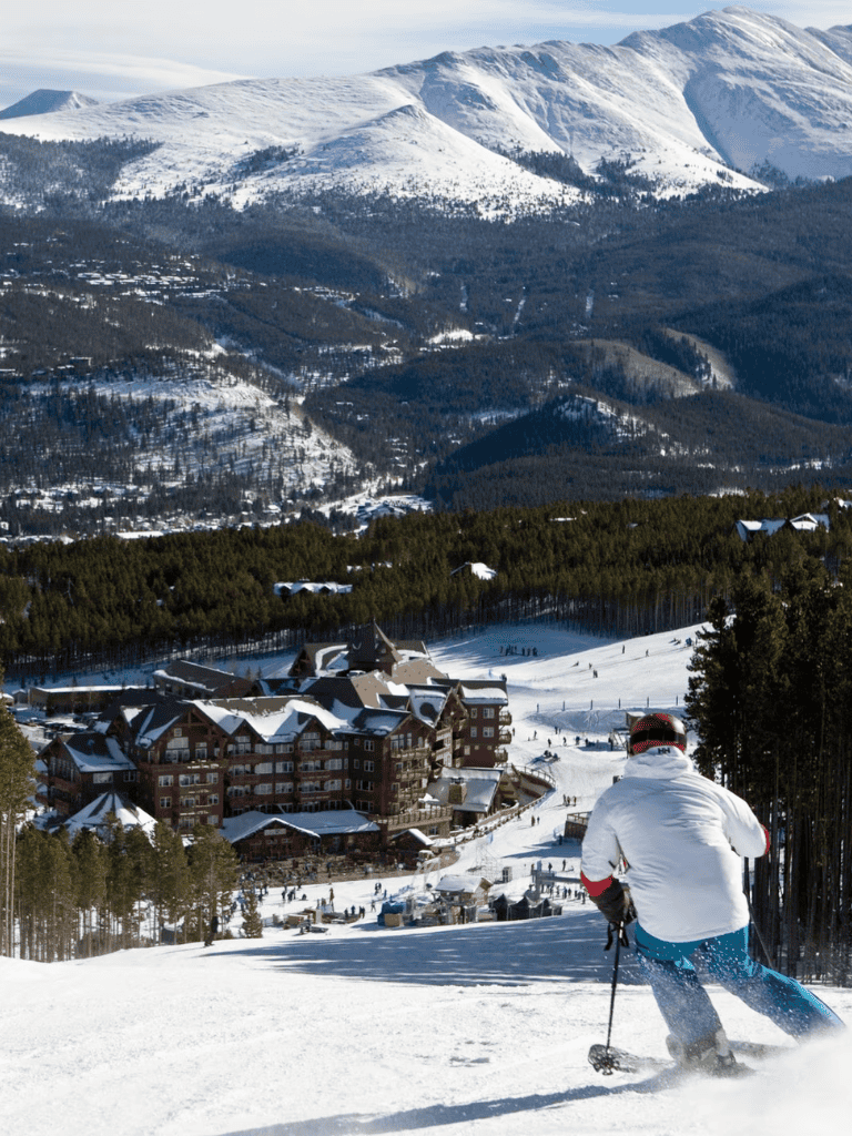 Skiing down snowy mountain with lodge and forest in background, scenic winter landscape.
