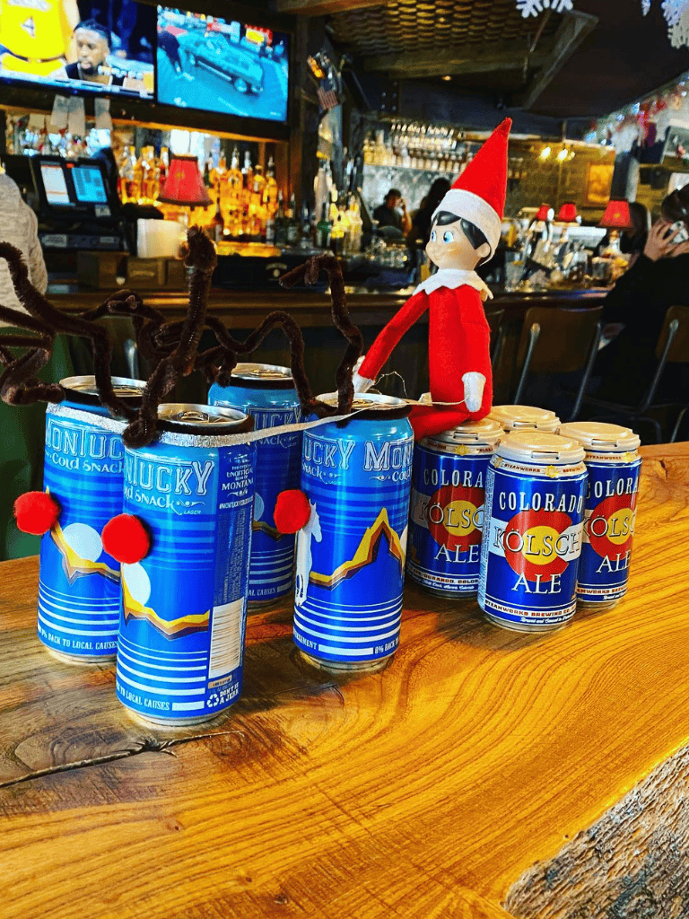Blue Chuck Montana beer cans with Santa elf decoration in a bar setting, perfect for holiday celebrations.