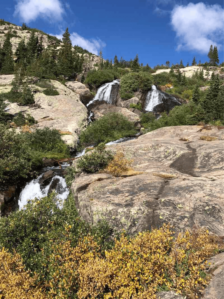 Serene mountain waterfall cascading over rocks with evergreen trees and blue sky, nature adventure scene.