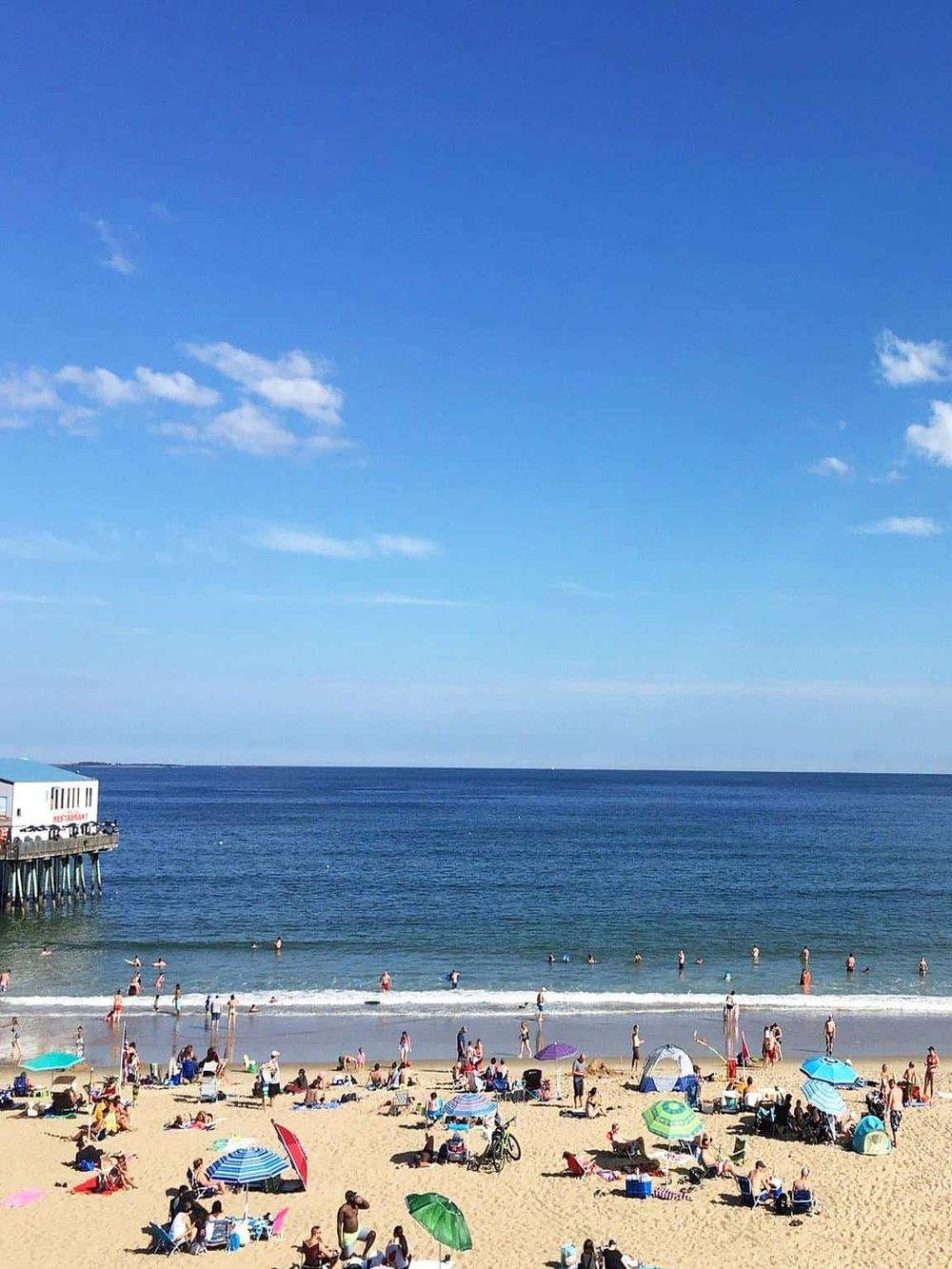 Vibrant beach scene with people enjoying sunny day, ocean views, colorful umbrellas, and pier, perfect for beach vacation and coastal relaxation.