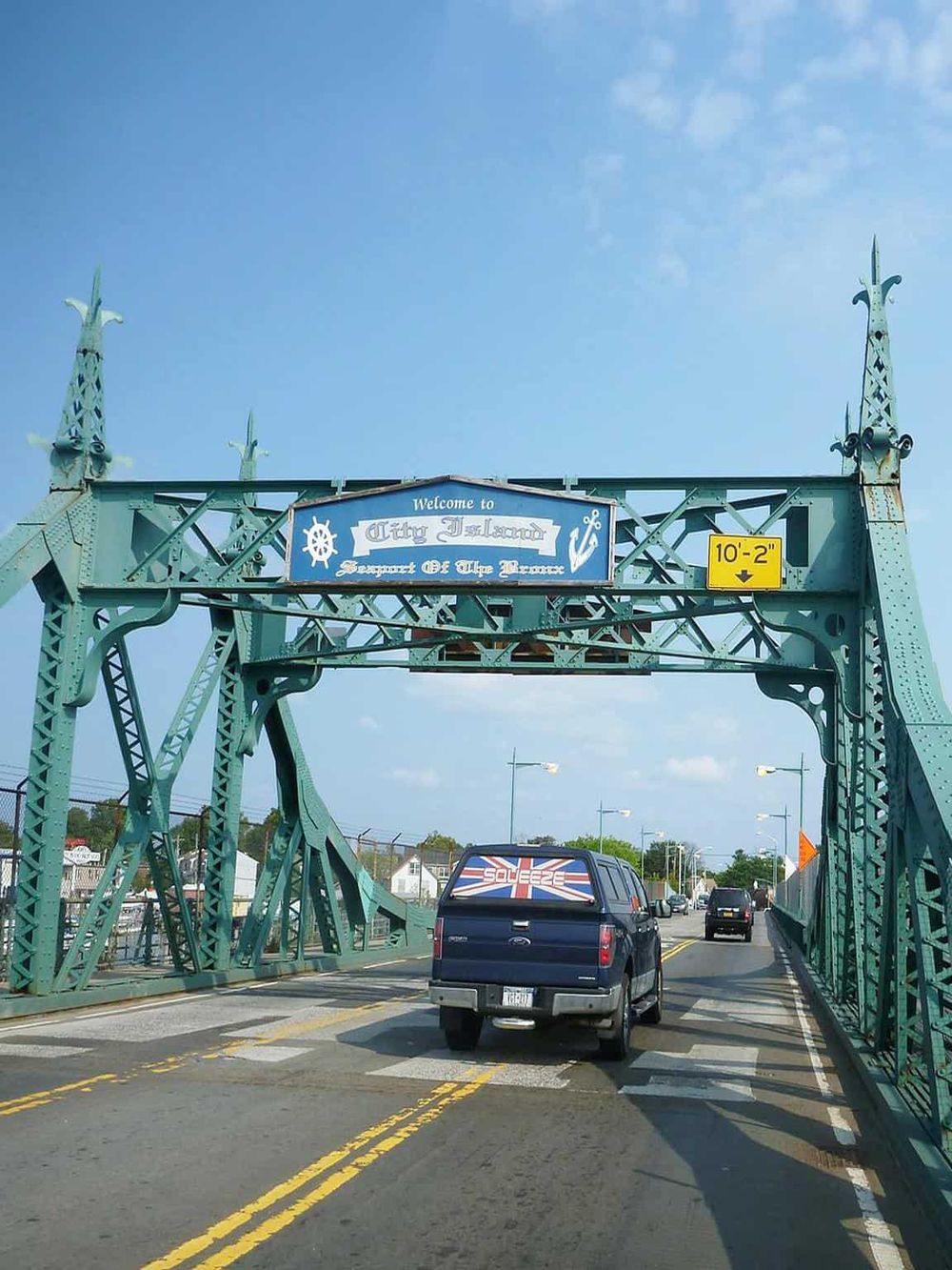 Bright green bridge entrance with "Welcome to City Island" sign, cars driving on a sunny day.