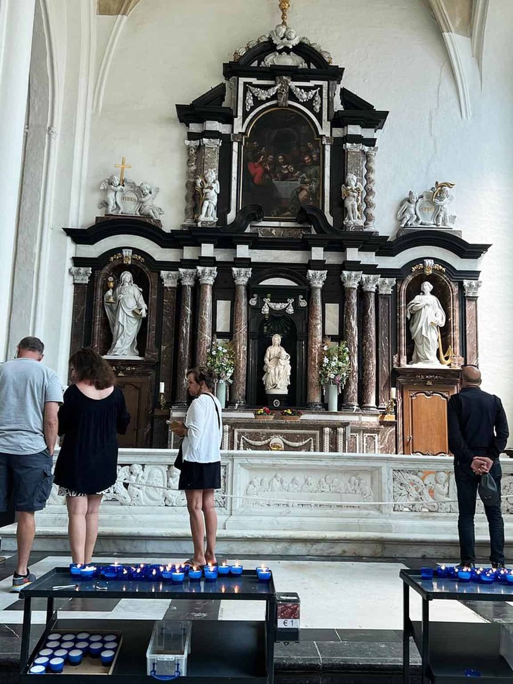 Shaded, ornate church altar with visitors lighting candles, intricate statues, and religious art, highlighting spiritual worship and faith practices.