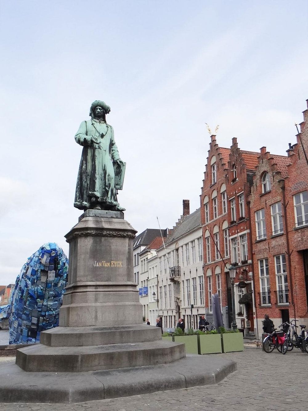 Bronze statue of Jan Van Eyck in historic European city square, surrounded by brick buildings and bicycles.