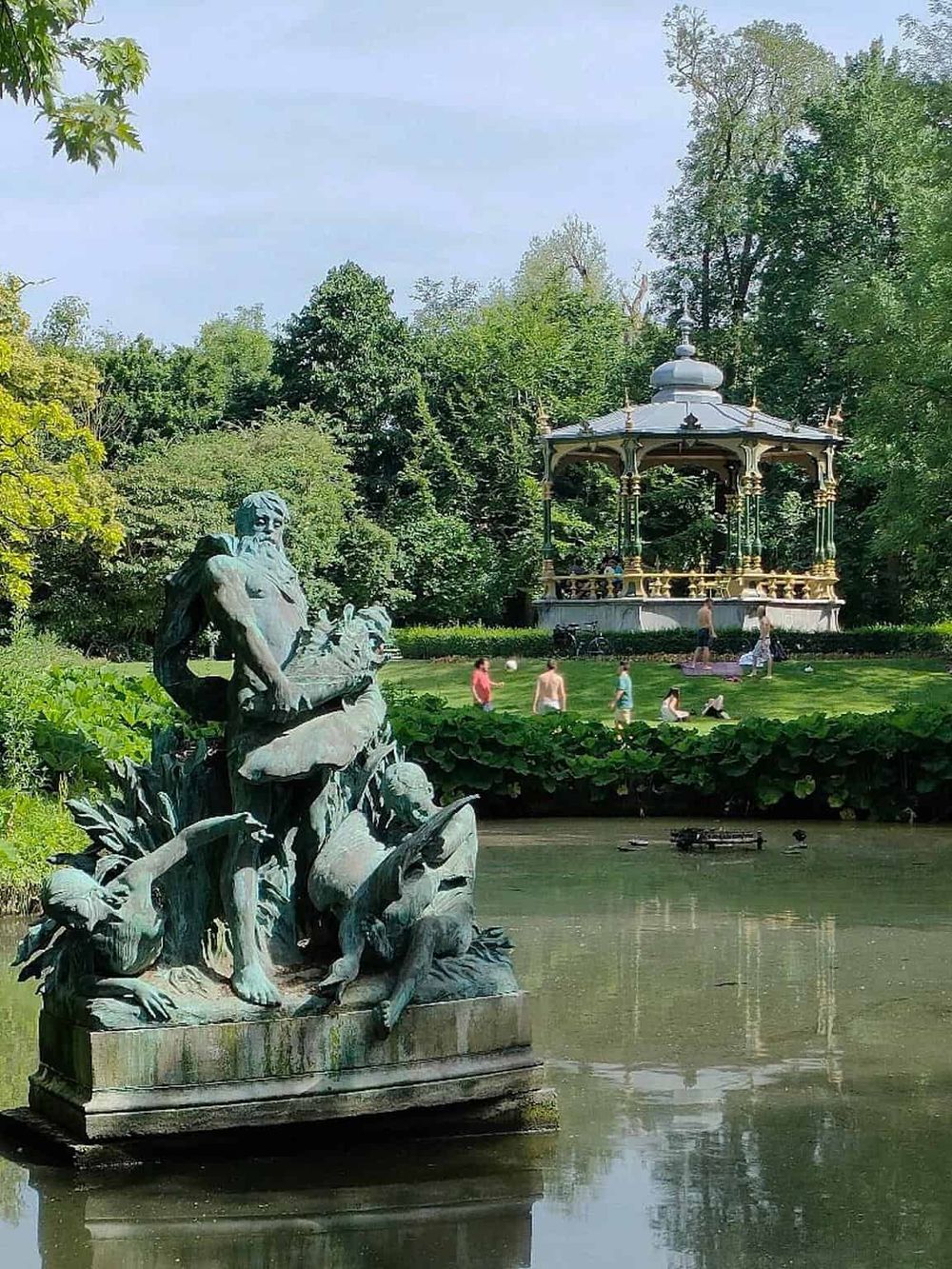 Ancient fountain sculpture in a lush park with a gazebo and people enjoying outdoor activities.
