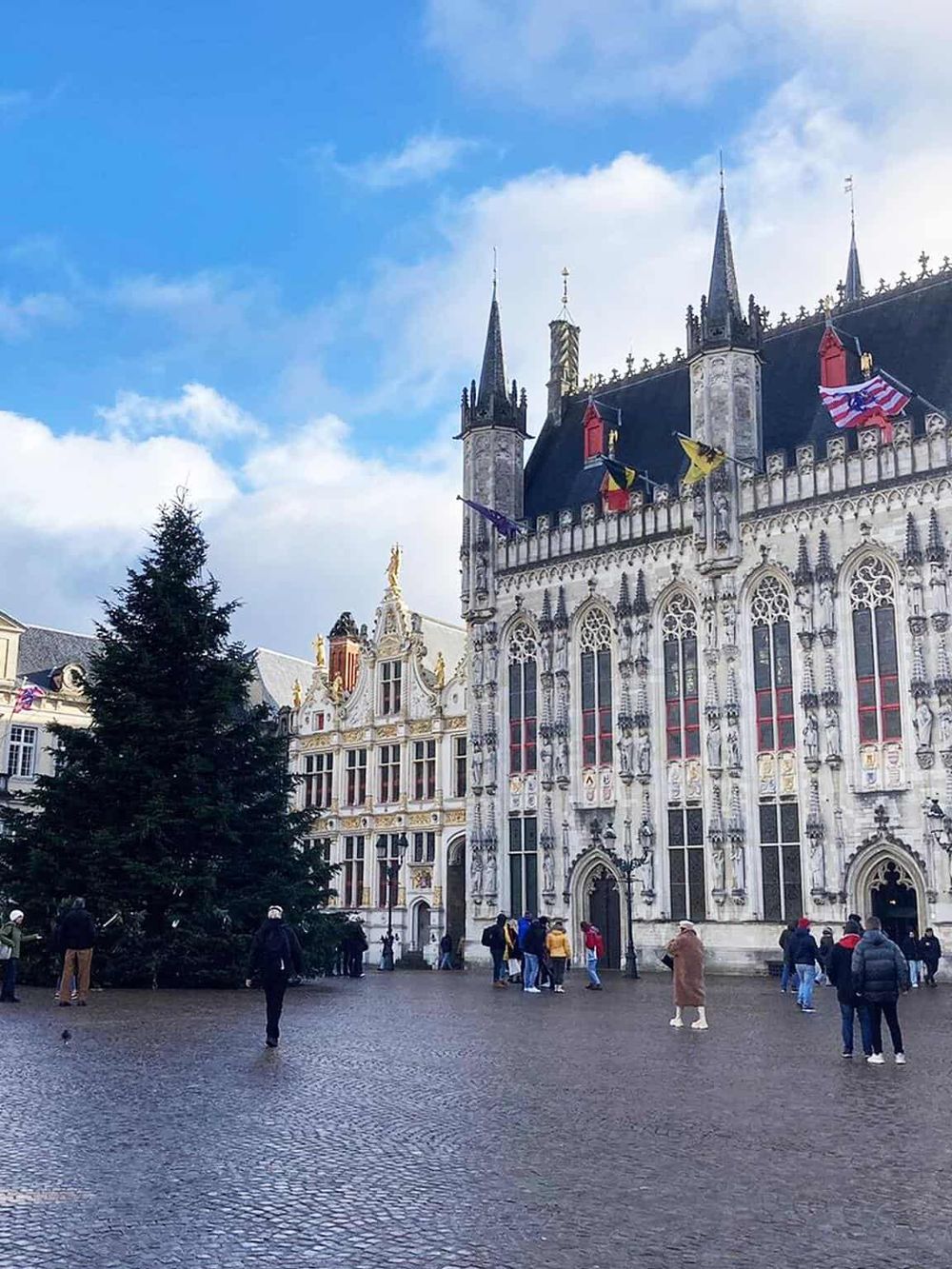 Colorful historic buildings in Bruges, Belgium, with a Christmas tree in the main square and tourists exploring.