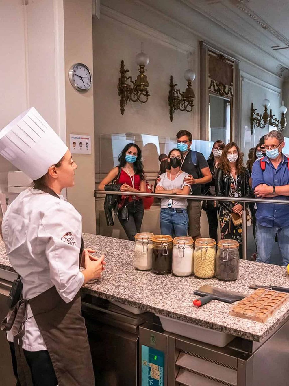 1. Pastry chef explaining dessert options to a group of mask-wearing visitors at a bakery.