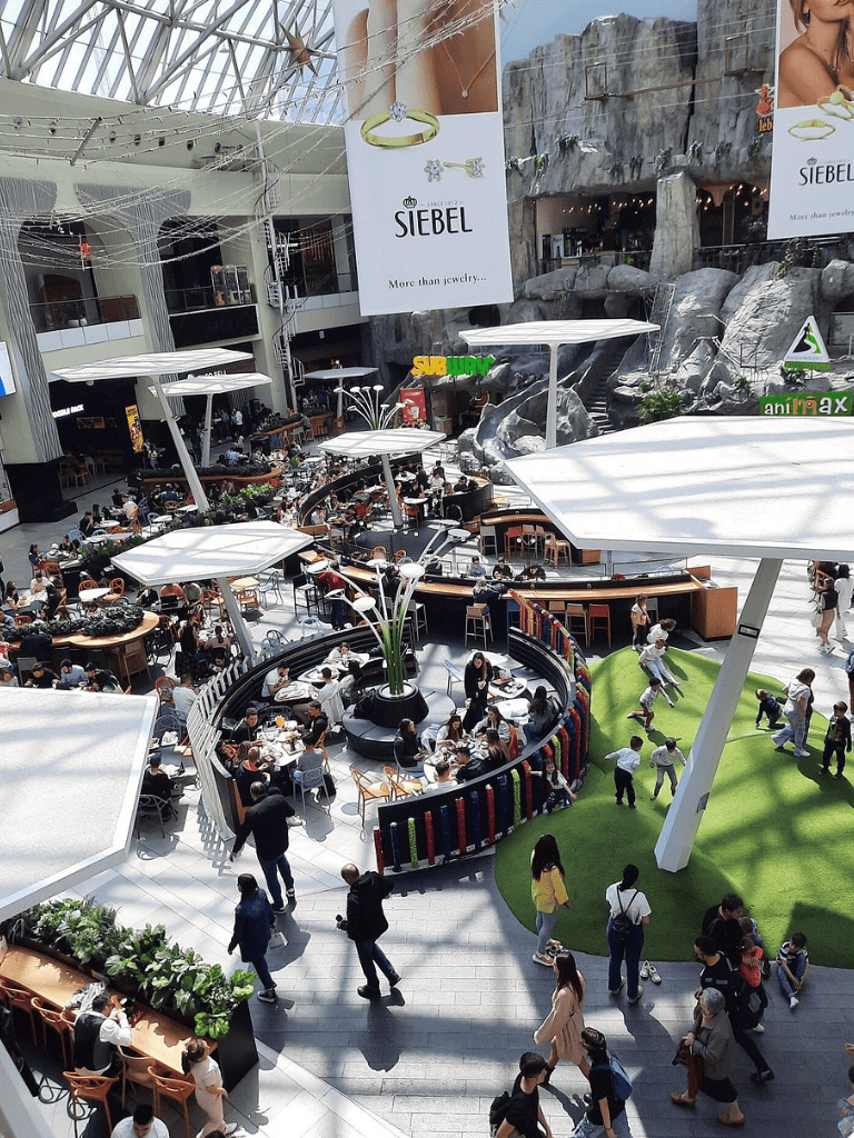 Indoor shopping mall atrium with dining area, children playing, and shoppers.