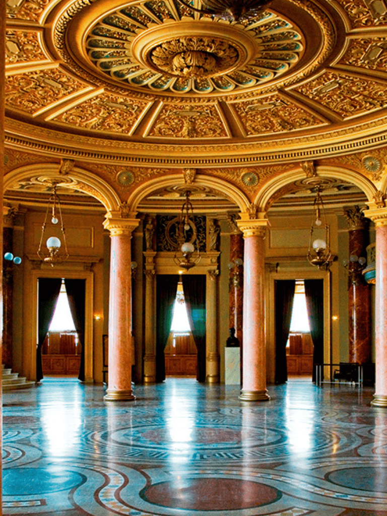 Opulent historic interior with ornate gold ceiling, marble columns, and elegant lighting fixtures.