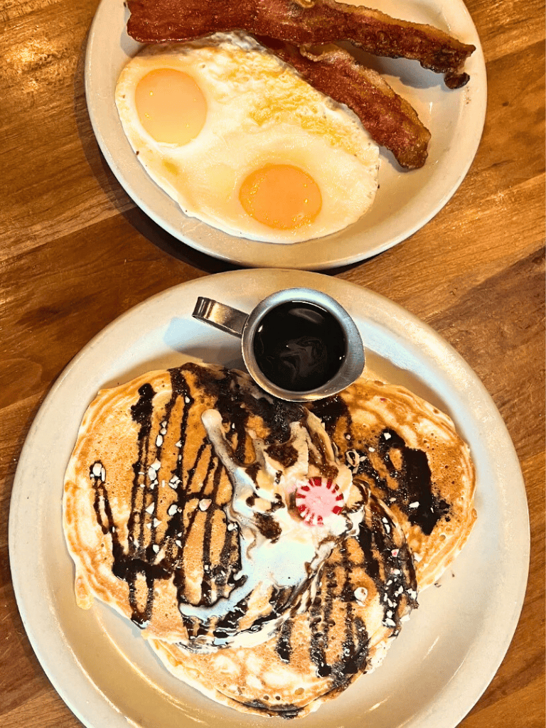 Fluffy pancakes with chocolate syrup, whipped cream, and a side of fried eggs with bacon on a wooden table.
