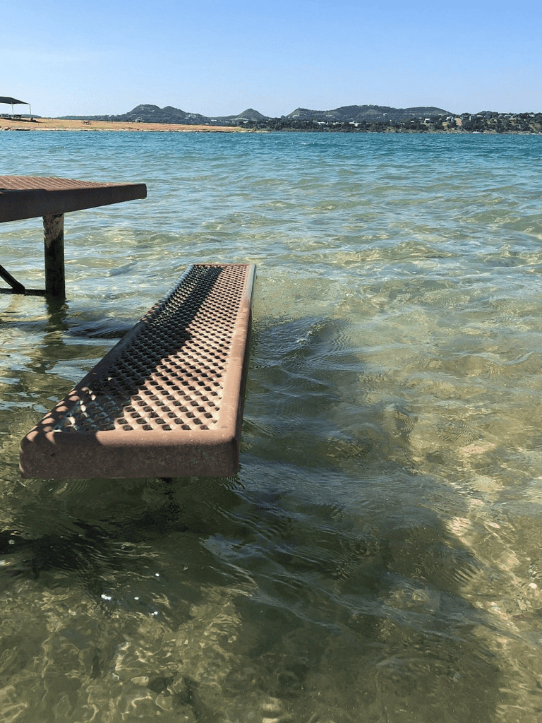 A close-up of a bench submerged in clear, shallow water at a beach shoreline with distant hills and a blue sky.