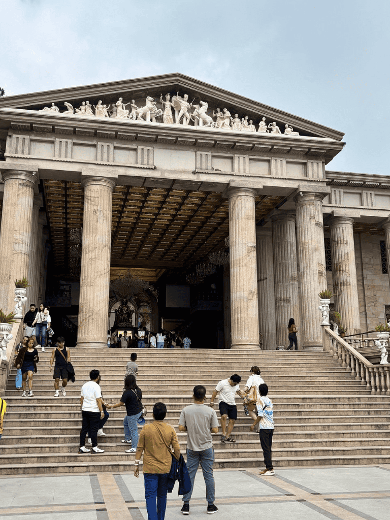 Ancient Greek-inspired building with steps and columns, visitors entering, Greek-style architecture.
