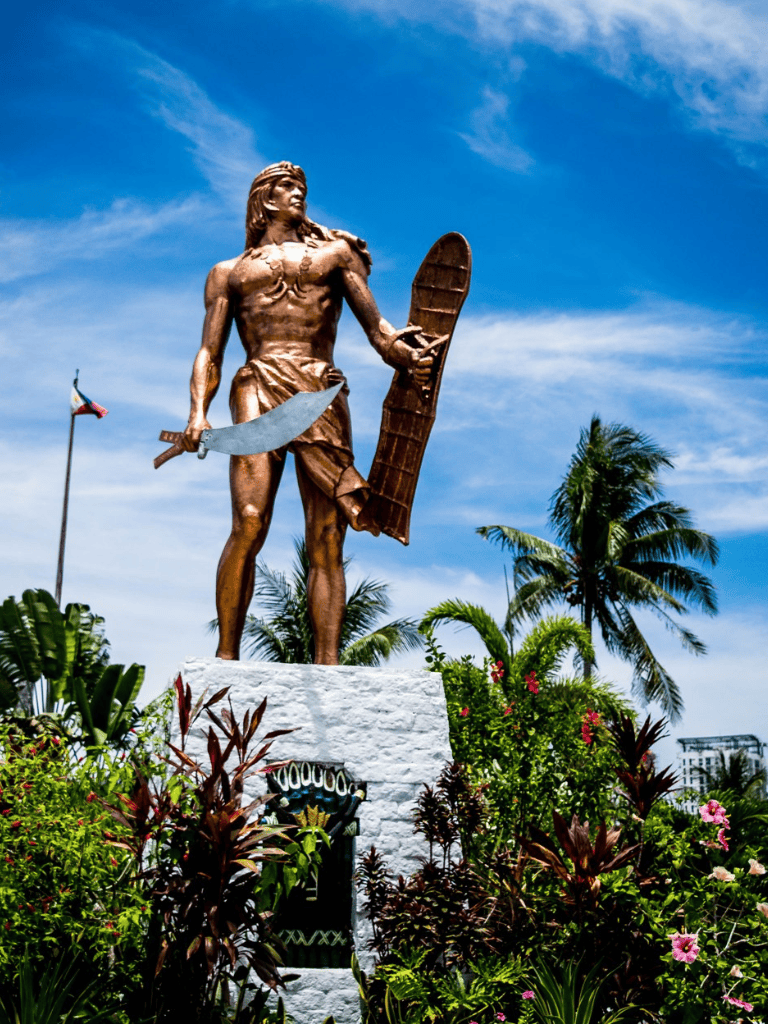 Bronze statue of a warrior with shield and sword in tropical garden with palm trees and blue sky.