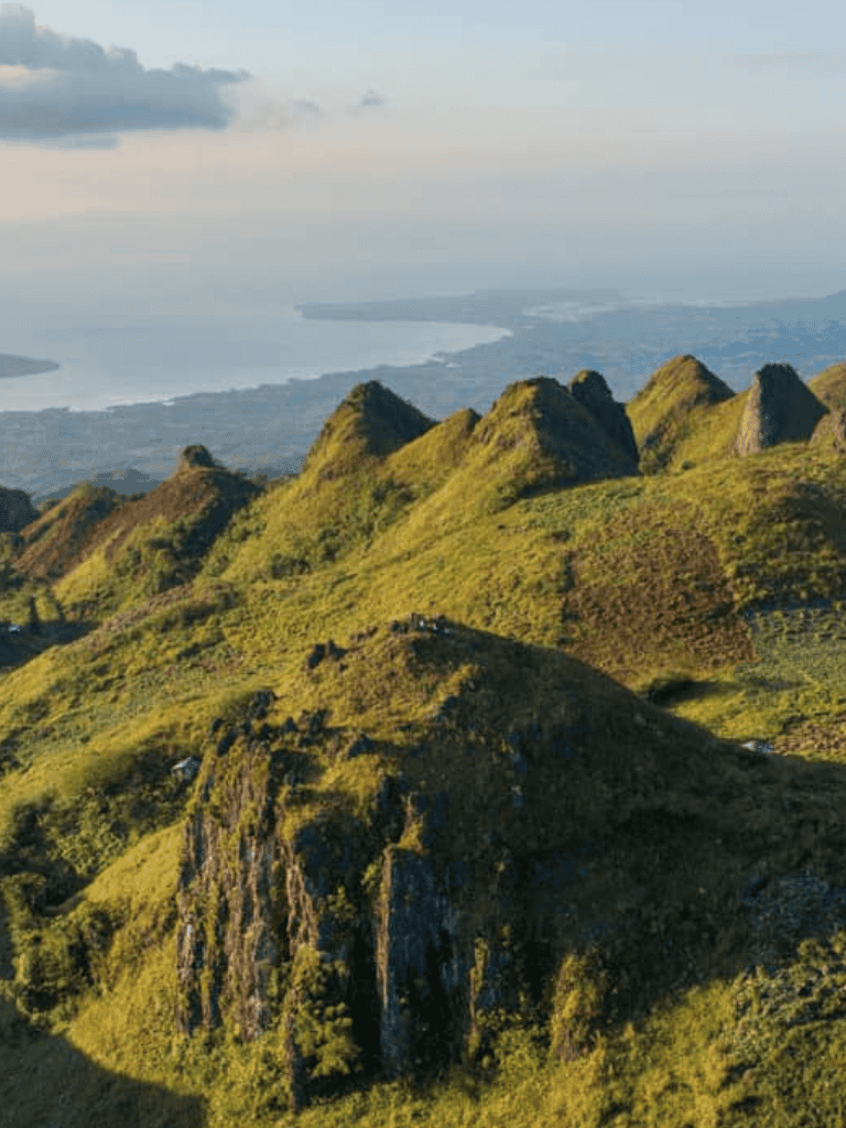 Vibrant green mountain landscape with lush hills and rocky outcroppings overlooking a distant body of water.