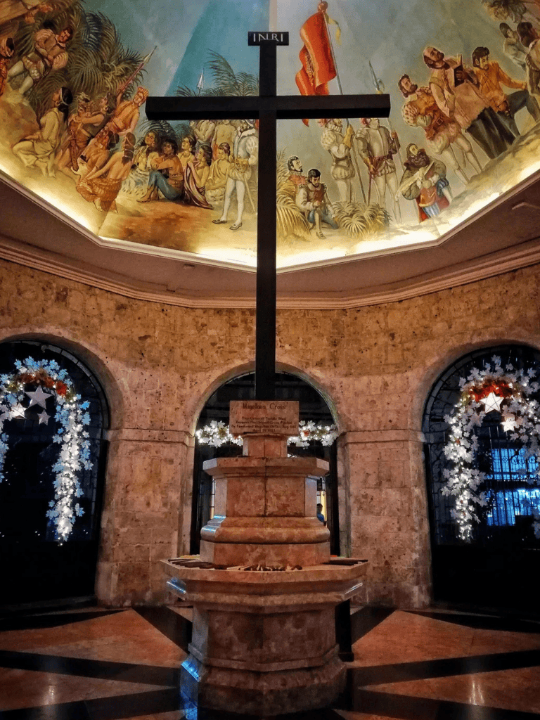 A religious cross with a painted ceiling featuring biblical scenes, situated in a church decorated with Christmas lights.