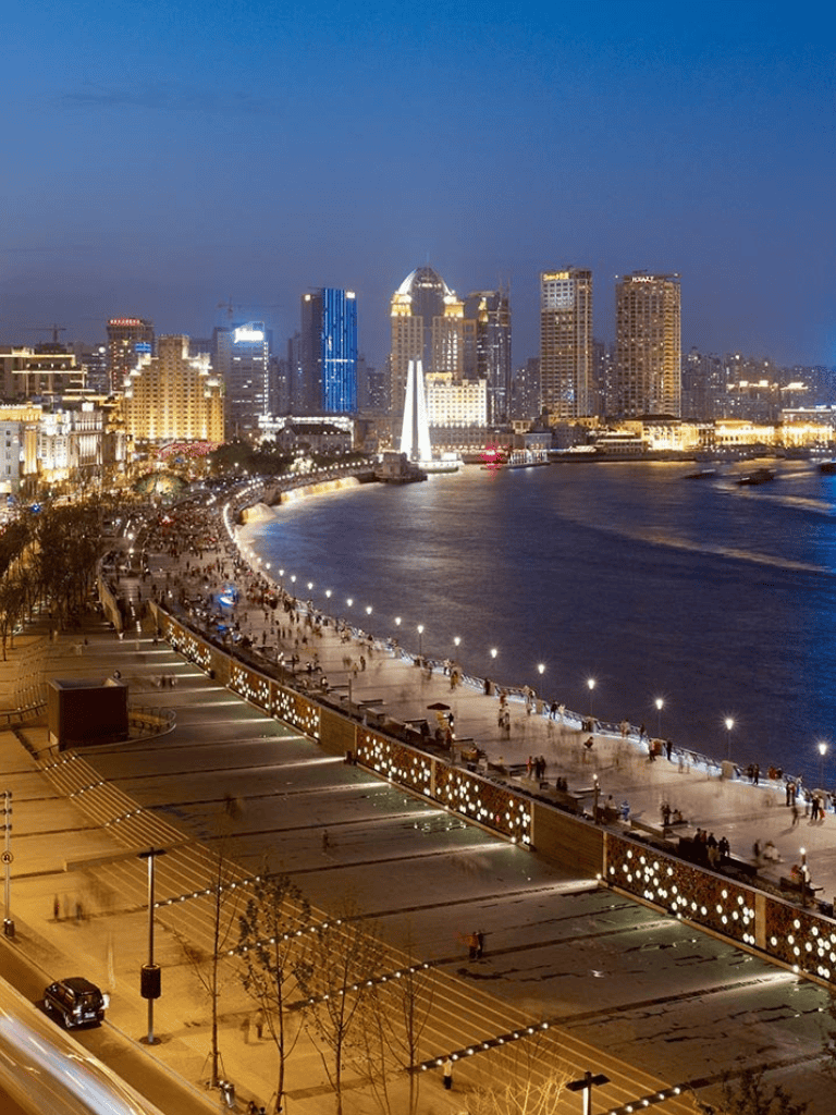 Night view of downtown city skyline along waterfront promenade with illuminated buildings and busy walkway, vibrant cityscape with scenic river.