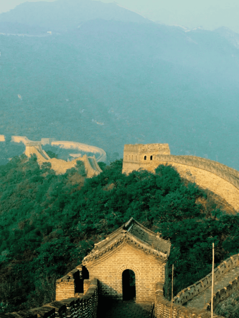 Ancient Great Wall of China winding through lush green mountains under a soft sky.