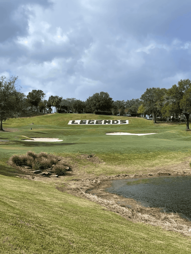 Vibrant golf course with "Legends" landscape design and scenic water hazard under cloudy sky.