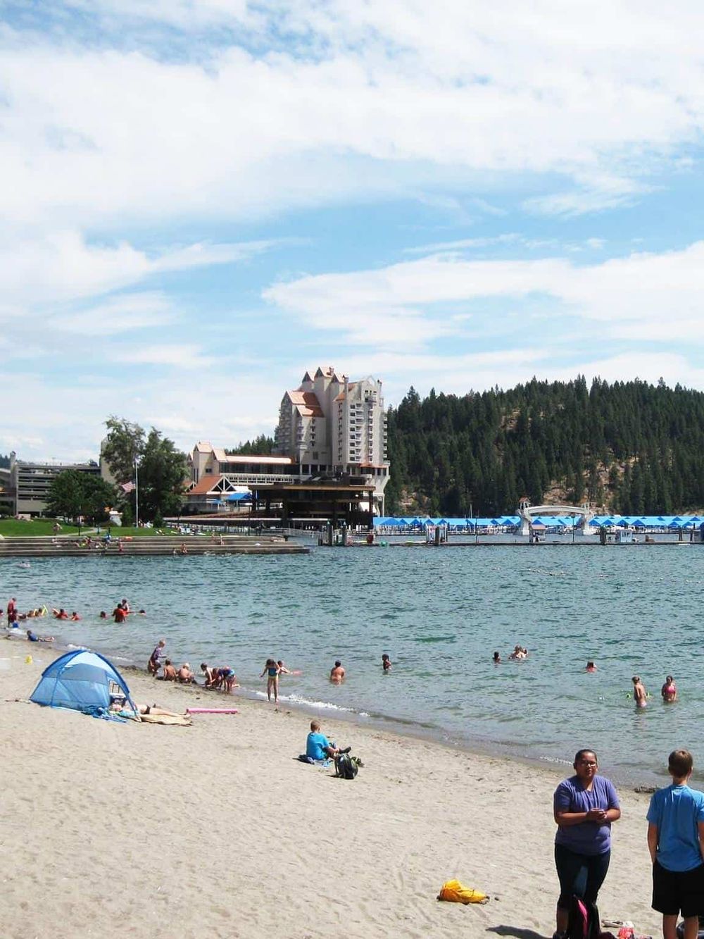 Scenic beach at Lake Tahoe with people swimming and relaxing, surrounded by trees and a resort building in the background.