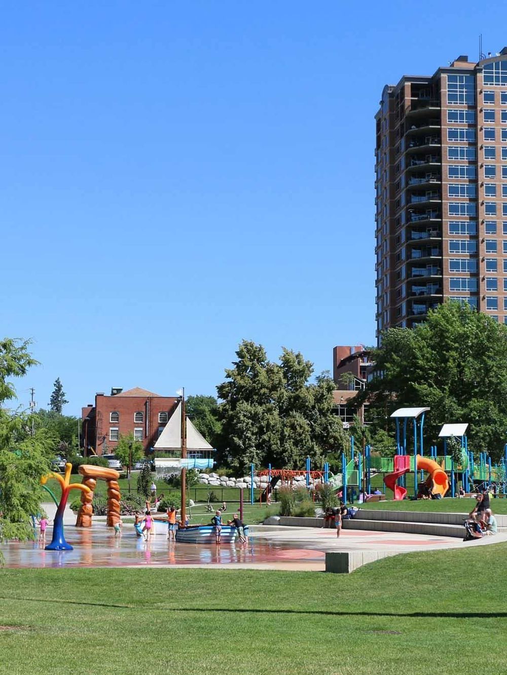 Vibrant urban park with playground, water features, and tall residential buildings under a clear blue sky.