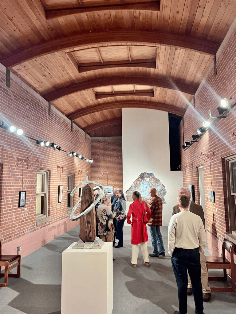Vibrant art gallery with brick walls, wooden ceiling, and contemporary sculptures and paintings.