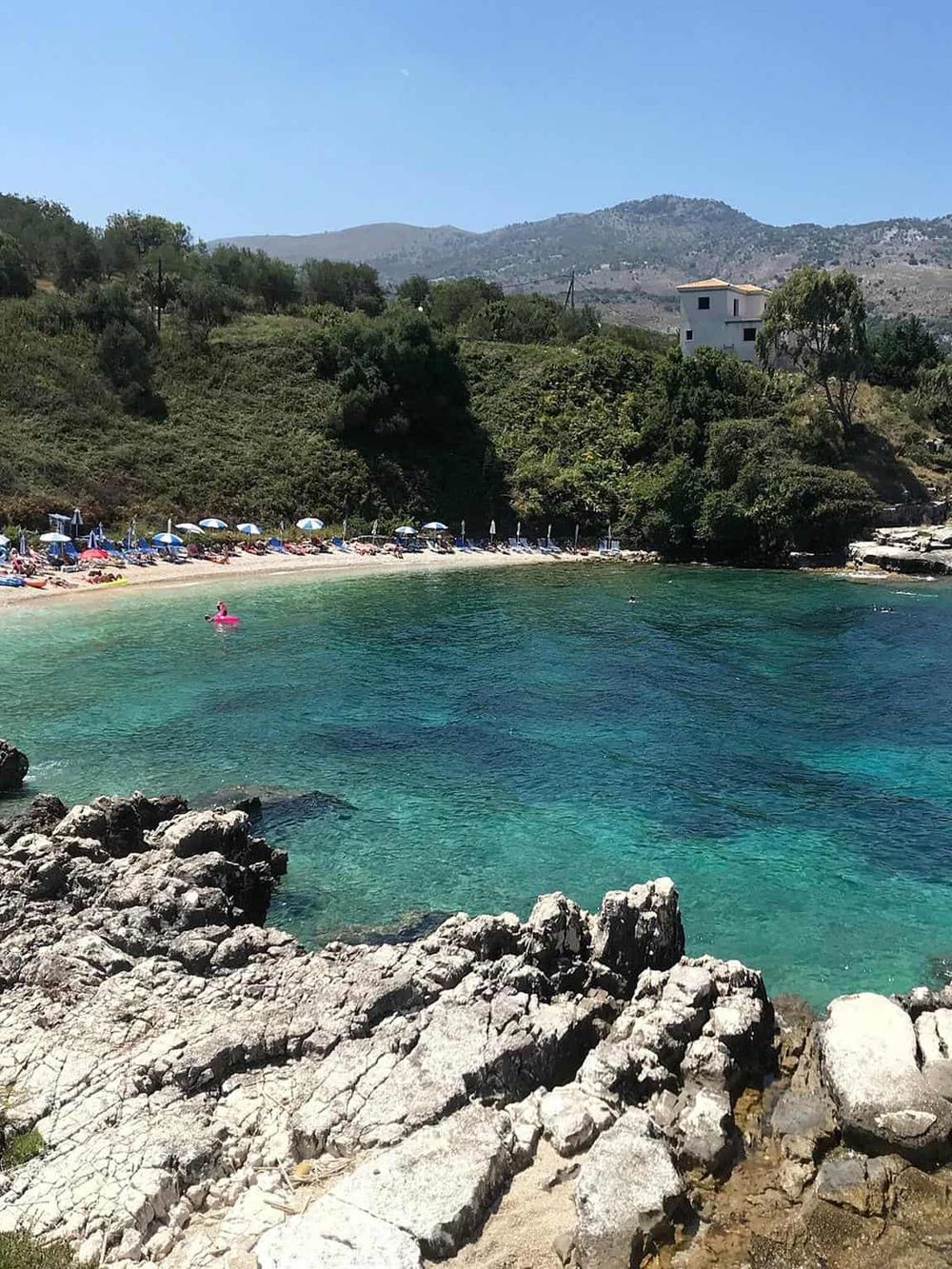 A scenic beach with turquoise water, surrounded by rocks, lush greenery, and hillside residences, perfect for travel and relaxation.