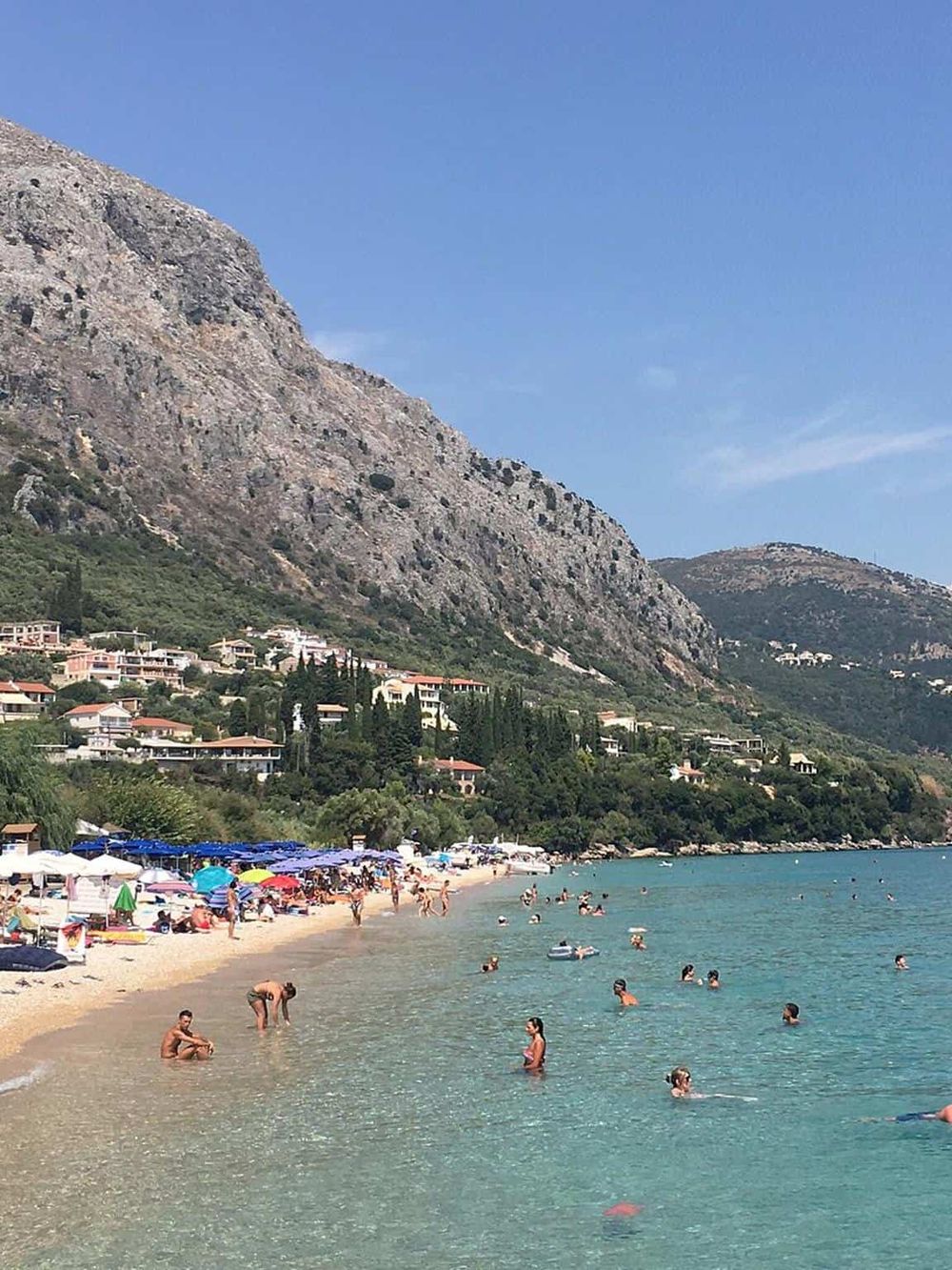 Beach with colorful umbrellas and people swimming, scenic mountains and hillside homes in the background.