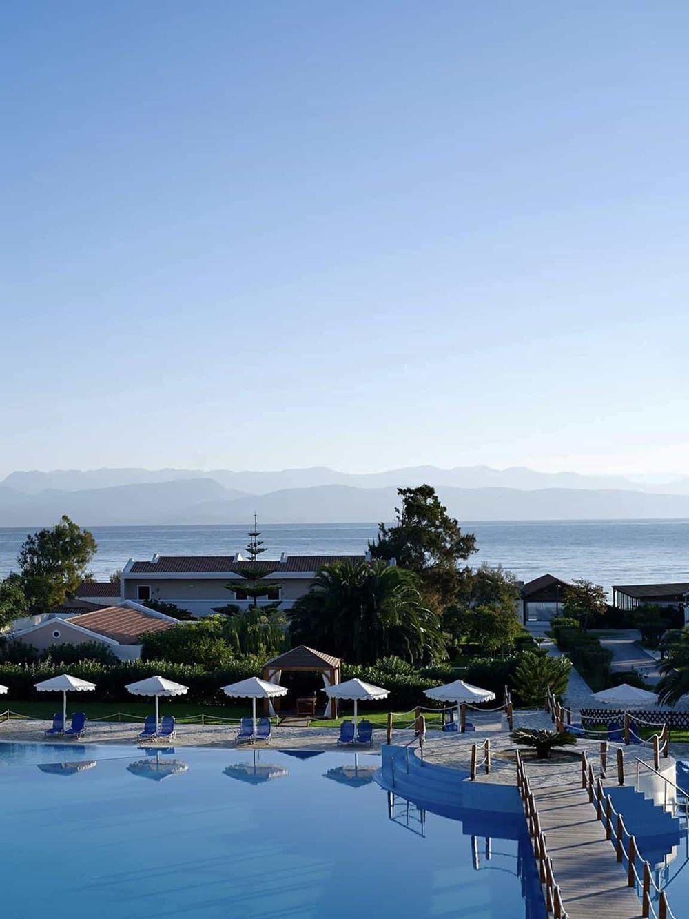 Stunning ocean view of a resort with a pool, umbrellas, lush greenery, and mountains in the background.