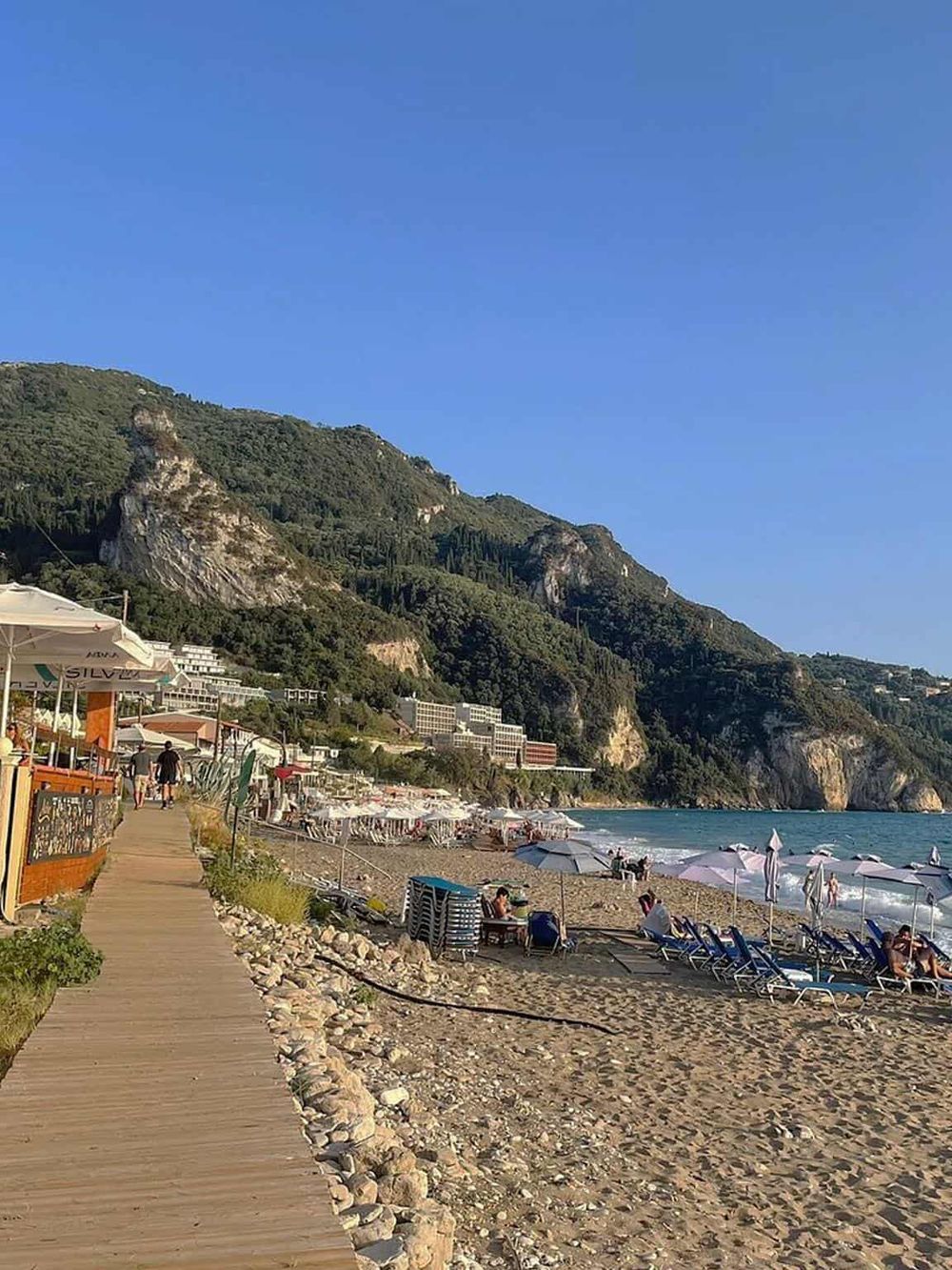 Scenic Mediterranean beach with sun loungers, umbrellas, and lush green cliffs in the background.