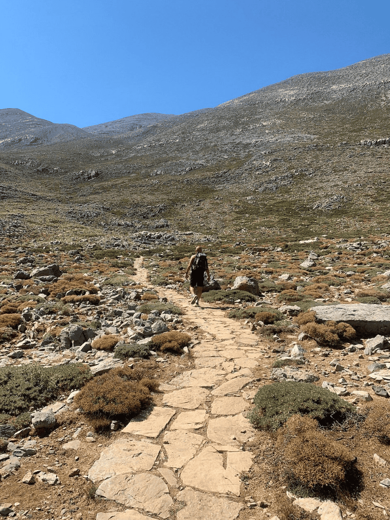 Scenic mountain trail for hiking with a solo hiker in a rocky, arid landscape under clear blue sky.