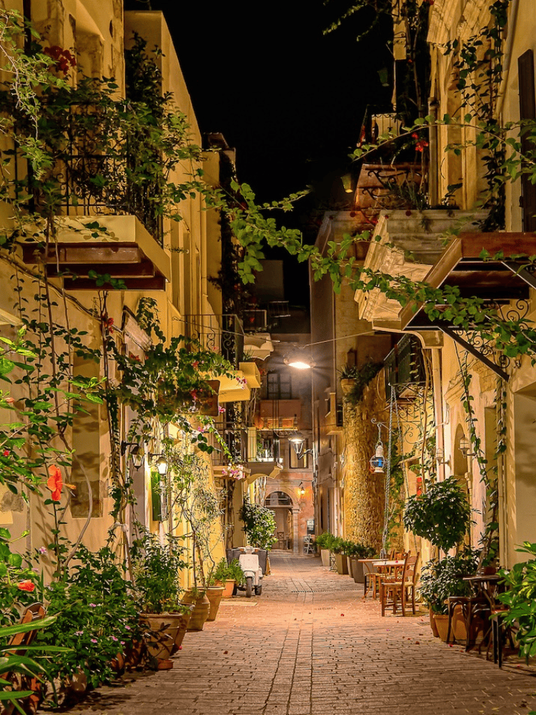 Charming night street scene with outdoor cafes, floral decorations, and warm lighting in a European-inspired neighborhood.