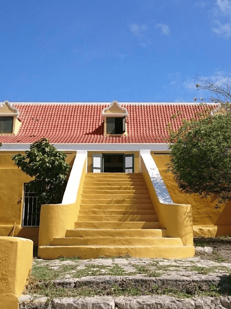 Bright yellow house with red roof, stairs leading to entrance, lush trees, clear blue sky, peaceful curb appeal.