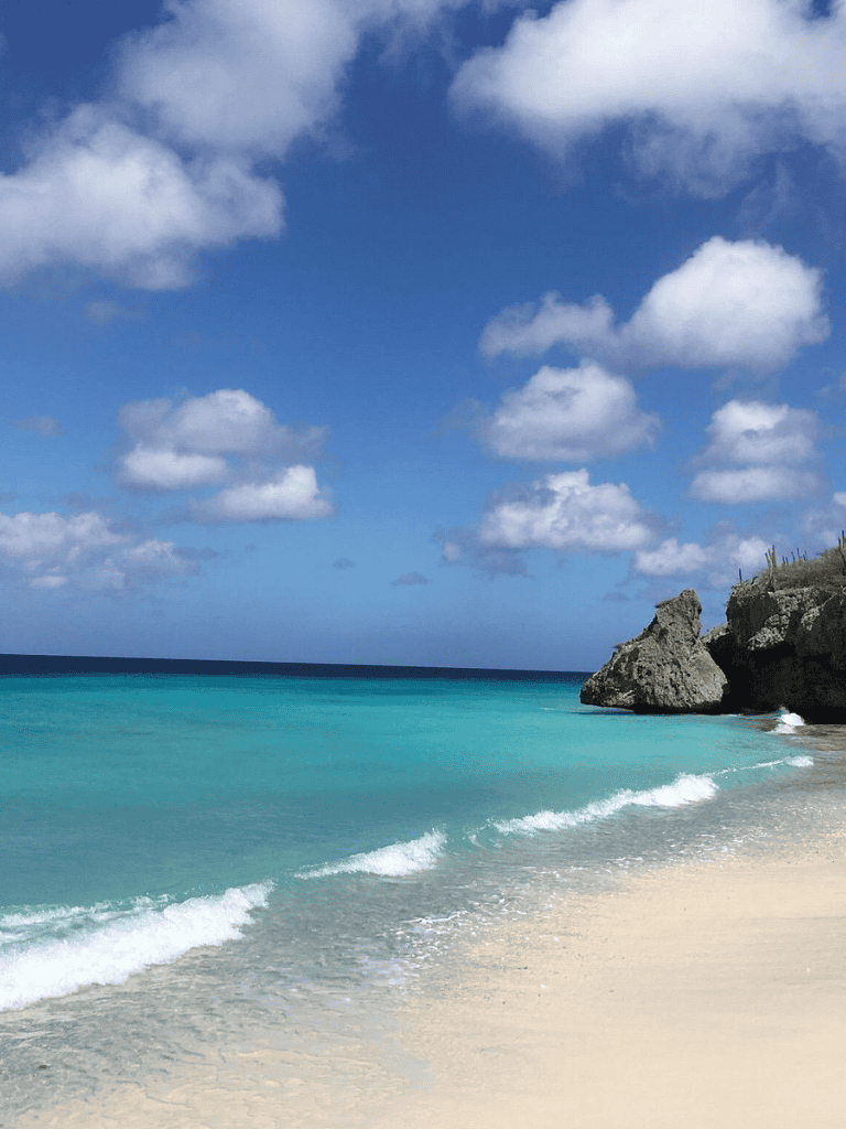 Pristine beach with turquoise water and rocky cliffs under a blue sky with fluffy clouds.