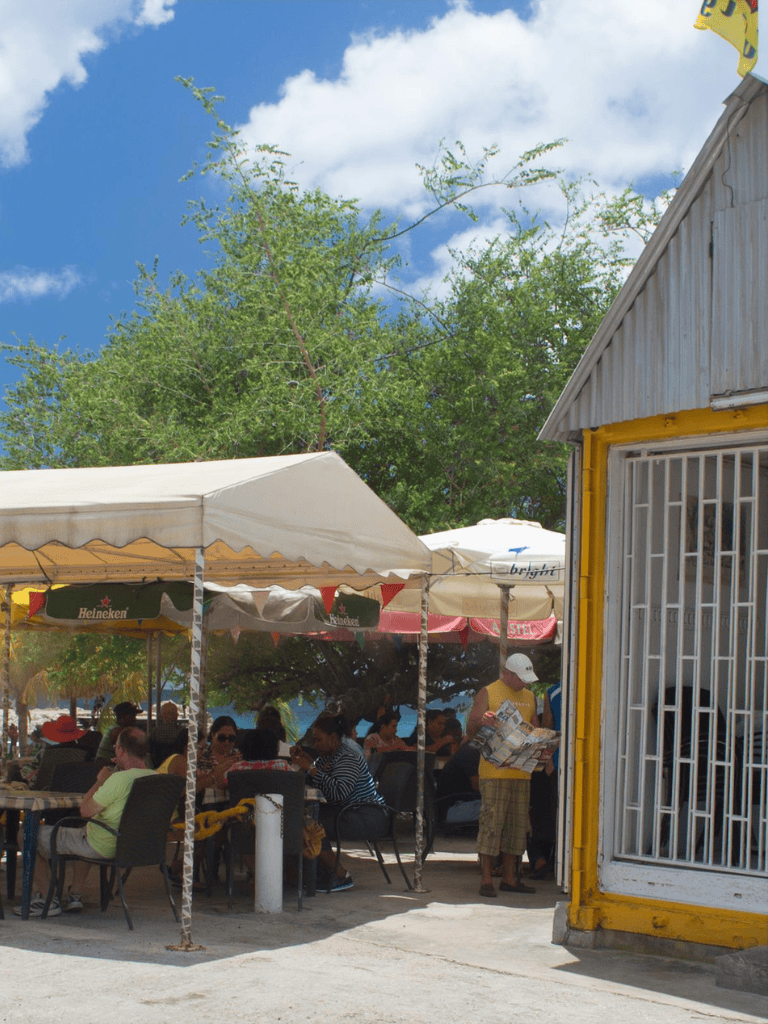 Outdoor restaurant with shaded seating and groups of people enjoying meals, sunny weather, lush green trees, colorful umbrellas, and a blue sky.