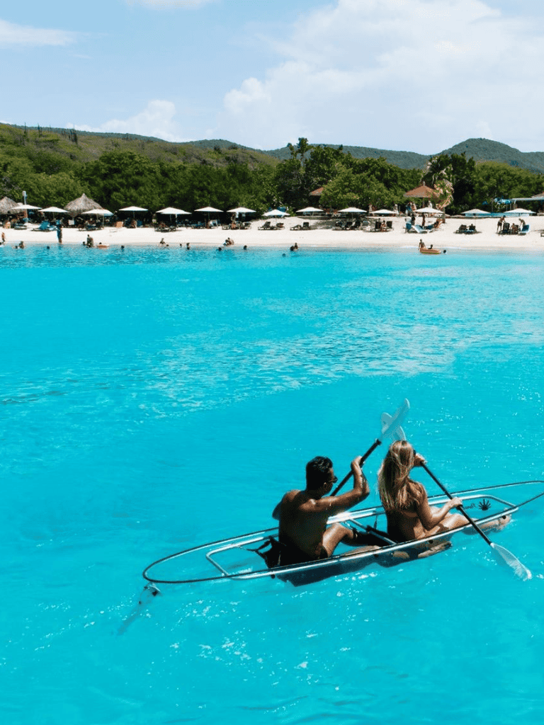 Pristine blue water with people kayaking near lush green hills and beach umbrellas — ideal for travel adventures.