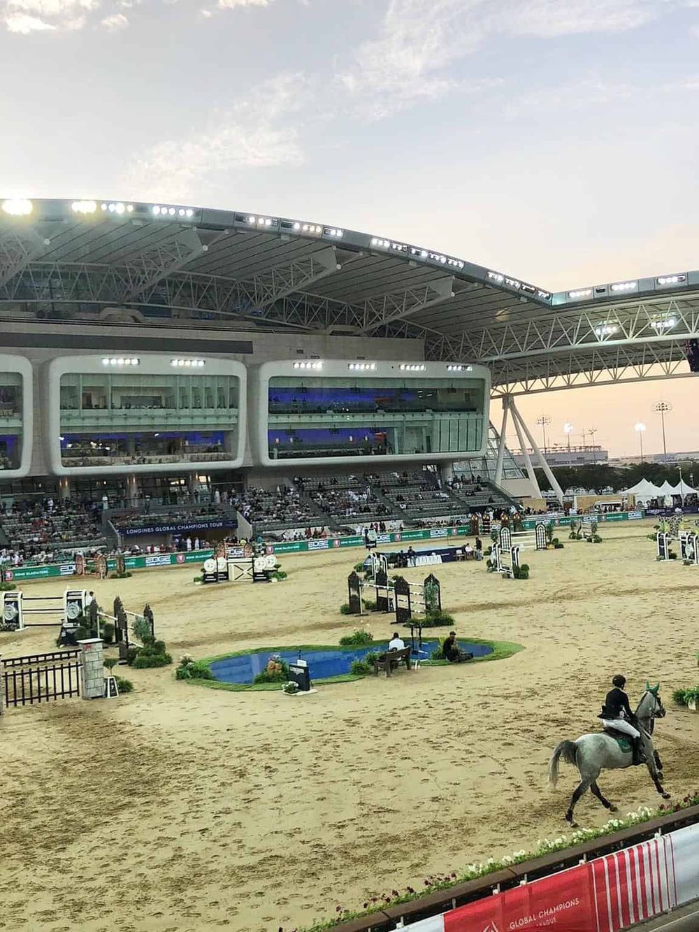 Modern indoor equestrian arena with spectators, show jumping course, and rider on horse during a competition.